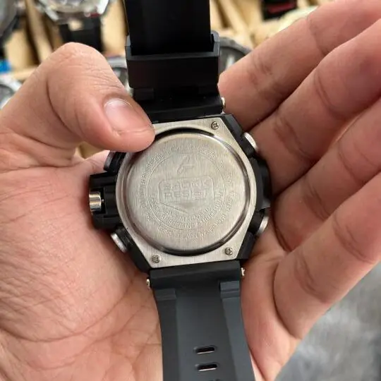 G-Sh0ck Steel Series analog-digital watch with silver stainless steel bezel and black rubber strap held in hand, watch collection visible in background