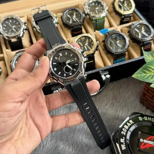 G-Sh0ck Steel Series analog-digital watch with silver stainless steel bezel and black rubber strap held in hand, watch collection visible in background