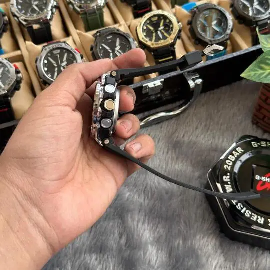 G-Sh0ck Steel Series analog-digital watch with silver stainless steel bezel and black rubber strap held in hand, watch collection visible in background