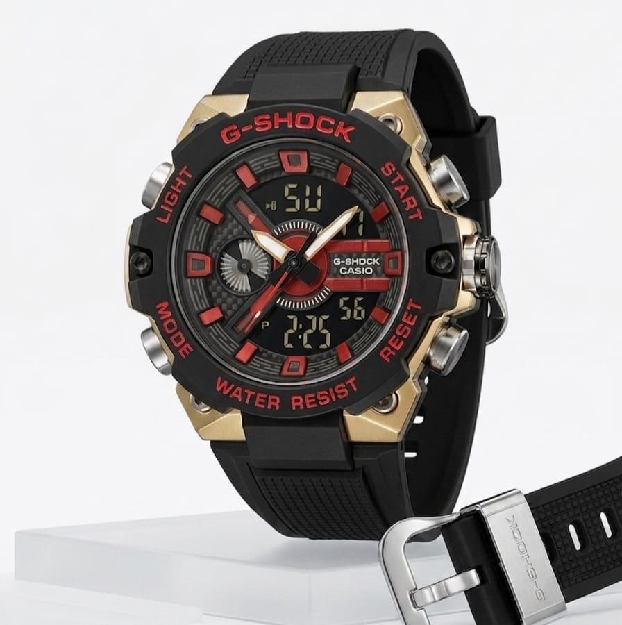 Casio G-Shock GST-B100 analog-digital watch with black carbon core case, gold metal bezel, red accent markers and black resin strap displayed on white surface with extra strap and buckle